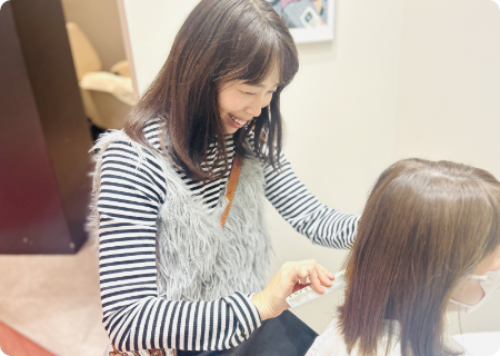 白いシャツを着た笑顔の女性美容師が、ツールベルトを下げ、右手にハサミを持ってカメラに笑顔を向けている様子